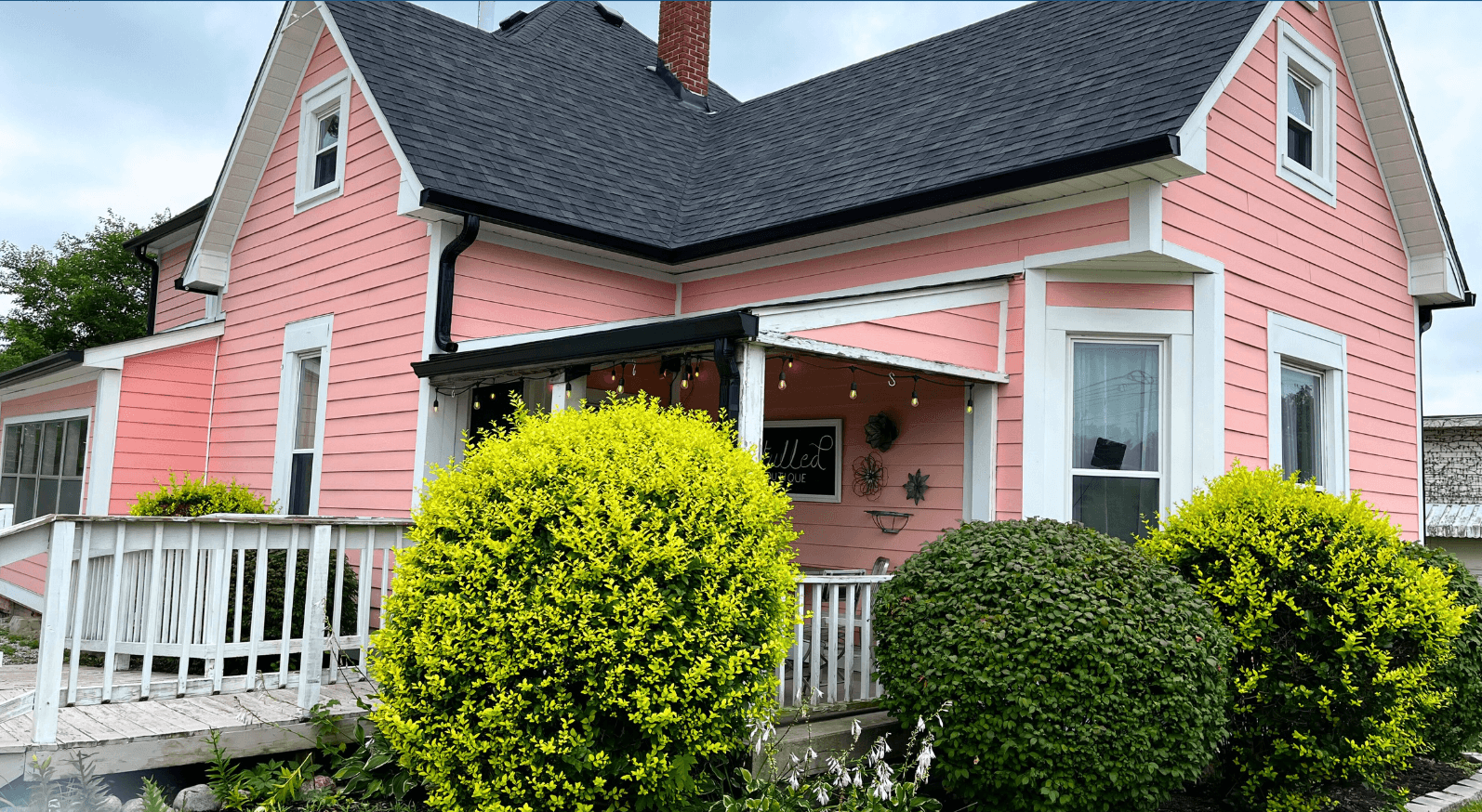 Exterior of a two-story historic home in Cumberland, Indiana, painted salmon pink with white trim and a dark gray gambrel roof. The building currently operates as Retulled boutique. A covered front porch is strung with Edison bulb lights and displays a black boutique sign. Rounded and trimmed shrubs line the front, and a white wooden deck ramp is visible on the left side.