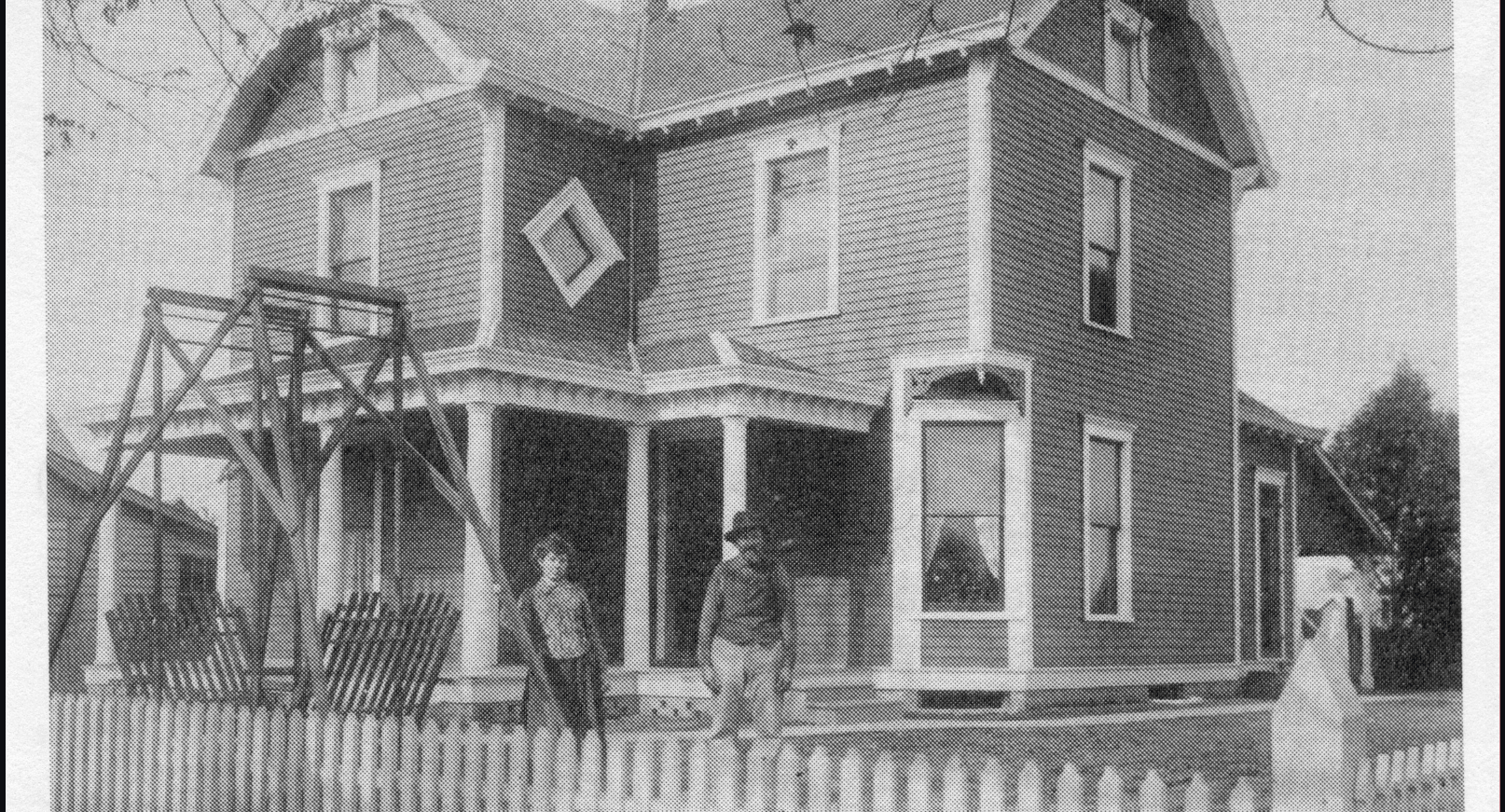 Black and white photograph, likely taken between 1890 and the early 1900s, of a two-story wood-frame house in Cumberland, Indiana. Two unidentified people stand on the covered front porch, which has decorative columns. A diamond-shaped attic vent is visible on the front gable. A tall wooden structure, possibly a windmill or water tower frame, stands to the left of the house.