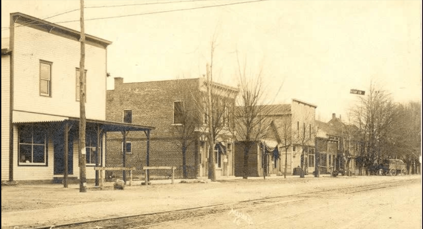 Sepia-toned historic photograph of Main Street in Cumberland, Indiana, era unknown. A row of two-story commercial buildings lines the street, including wood-frame storefronts on the left and brick buildings further down. Utility poles with overhead wires are visible, bare trees line the sidewalk, and horse-drawn wagons appear in the distance. A streetcar track runs along the unpaved road.